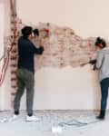Two people removing plaster from a brick wall in a room under renovation.