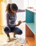 Woman painting the inside of a dresser aqua blue. She is in an empty room except for a ladder. There is a lot of light coming in.