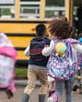 A multi-ethnic group of elementary age children are getting on a school bus. The kids' backs are to the camera. They are running towards the school bus which is parked with its door open. It's a rainy day and the kids are wearing jackets, rain boots and backpacks.