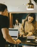 Woman signing papers at a hotel reception desk with two laptops and a gold pendant light above.