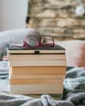pile of books with glasses and a laptop on the bed at home