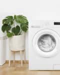 Washing machine in laundry room with potted plant, wicker basket, and shelf holding detergent and folded towels.