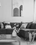 Grayscale photo of a square, wooden table standing in front of a brown, suede sofa with decorative cushions in cozy living room interior
