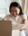 Woman sitting on sofa in living room; opens received delivered parcel and feels happy.
