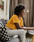 A young woman is using a laptop and working in the living room while smiling and enjoying in a cozy environment.