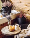 Cute autumn home decor arrangement. Tiny wooden cabin balcony with heather flowers in pot, lavender in bottle vase, candlelight flame, soft beige plaid waiting on comfortable garden furniture chair