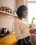 Woman standing in the kitchen holding a cup of coffee and texting on her cell phone. Woman using a mobile phone at home.