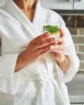 Cropped view of unknown woman in white waffle bathrobe holding a glass with lemon and mint water. Concepts of healthy lifestyle and diet.
