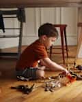 A young child in an orange shirt plays with various toy animals on a wooden floor under a table.