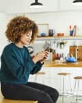 Young woman surfing social media at home. Beautiful female is using smartphone while sitting on table. She is wearing casuals.