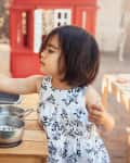 A toddler girl plays with her outdoor kitchen in her backyard on a warm, sunny day. There are pots and pans and kitchen tools and a table and chairs in the background and also a red playhouse.