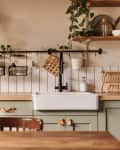 Empty kitchen island with marble surface in foreground, green vintage countertop with drawers and pendant lights hanging above, lots of flowers in jars, blurred background.