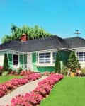 1950's green house with white trim with green lawn, pink azaleas. Mother and child outside with a large orange bucket