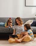 Beautiful woman with her kids sitting on the floor and using digital tablet for playing games, watching movies and relaxing at home. Her son is cuddled with her, while the daughter is lying on the sofa next to them. They are all smiling and looking down at the screen.