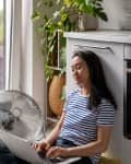 Woman typing on laptop while cooling by floor fan at home.