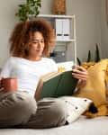 Young multiracial woman reading a book at home, drinking tea sitting on the sofa in cozy living room. Lifestyle concept.