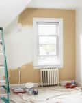 Room with a ladder, paint supplies, and partially painted beige wall near a window and radiator.