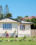 Single-story cream house with a metal roof, front steps, and a small garden, surrounded by trees and a wooden fence.