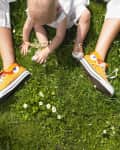 shot from above, a baby sitting inbetween someone's legs on the grass. grass has little white flowers. baby is wearing white clothes and orange hi-top sneakers. you only see the adult's legs and they are wearing orange hi-top sneakers as well.