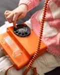 A teenage girl sits in a pink heart-patterned sweater, using a bright orange landline phone. The cozy bedroom setting conveys retro technology use and communication.