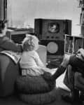 Family watching a small television in a living room, with a child sitting on cushions and a woman holding a magazine.