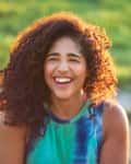 Smiling woman with curly brown hair wearing tie dyed tank top with green grass behind her