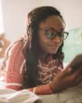 Photo of a young woman using her digital tablet while resting on the bed in her bedroom