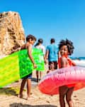 Photo of a young family of four enjoying a day at the beach; children are carrying inflatable beach toys and youngest daughter is bringing up the rear as she looks at the camera with a big smile.
