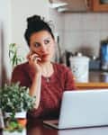 woman sitting in kitchen with a laptop. She is on the phone and has a blue coffee mug