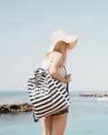 Woman in a sun hat and striped bag standing by the sea, with a boat in the distance.