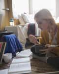 Woman sitting on floor eating from a pot, surrounded by books and records in a cozy, cluttered room.