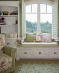 Cozy reading nook with floral cushions, green armchair, and white shutters overlooking a scenic view.