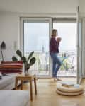 Living room with a woman holding a mug, standing by open balcony doors, plants, and a stack of books on the floor.