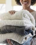 Woman holding a stack of folded knit sweaters in neutral tones.
