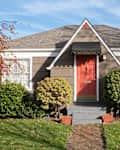 Charming brown house with a red front door, surrounded by green shrubs and a brick pathway.