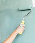 Two hands painting a wall with teal paint using rollers.