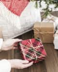Hands placing a plaid-wrapped gift under a Christmas tree with other presents and a red ornament.