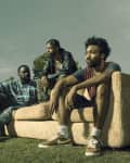 Three men sitting on a beige sofa in a grassy park, surrounded by trees under a cloudy sky.