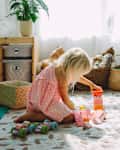 A young child in a pink dress plays on a soft rug, stacking colorful blocks beside toy baskets and a potted plant.
