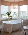 Freestanding bathtub in a bay window bathroom with a modern chair, plant, and chandelier.