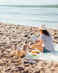 Woman in striped shirt and hat reading on a beach blanket with a straw bag, flip-flops, and snacks nearby.