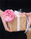 Person holding a gift wrapped in brown paper with a pink ribbon and a pink flower on top.