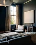 Cozy living room with dark walls, large window, gray sofa, striped ottoman, and a bar cart on a metal shelf.