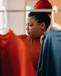 woman with hair up in braids is holding up a shirt she pulled off a clothing rack and examining it. There is a window letting in daylight in the background
