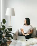 Woman sitting on a cream sofa with a book, next to a large plant and floor lamp, looking out the window.