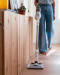 person in jeans vacuuming laminate floor, window and tall plant behind her