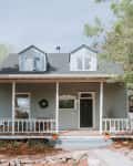 Two-story gray house with a porch, white railing, and dormer windows, surrounded by trees and a garden.