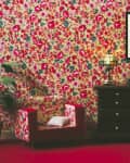 Floral wallpapered room with matching armchair, potted plant, dark dresser, stained glass lamp, and framed floral painting.