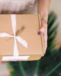 Hands holding a gift in a cardboard box with pink ribbon and tiny sprig of flowers