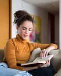 Woman in a mustard turtleneck reading a book on a beige sofa in a modern living room.